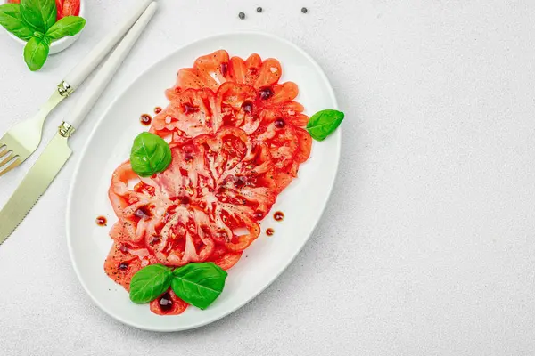 Dilimlenmiş Coeur de Boeuf domatesi, balzamik damlalar ve oval tabakta fesleğen yaprakları. Vejetaryen dostu, aile yadigarı, taş beton arka planda bir çiftlikten bir masaya estetik.