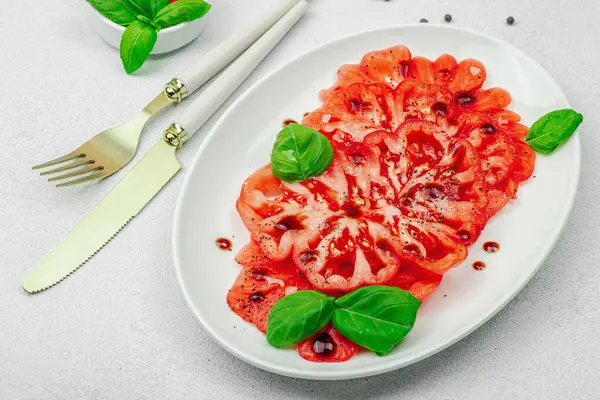 Dilimlenmiş Coeur de Boeuf domatesi, balzamik damlalar ve oval tabakta fesleğen yaprakları. Vejetaryen dostu, aile yadigarı, taş beton arka planda bir çiftlikten bir masaya estetik.