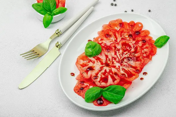 Dilimlenmiş Coeur de Boeuf domatesi, balzamik damlalar ve oval tabakta fesleğen yaprakları. Vejetaryen dostu, aile yadigarı, taş beton arka planda bir çiftlikten bir masaya estetik.