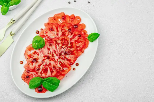 Dilimlenmiş Coeur de Boeuf domatesi, balzamik damlalar ve oval tabakta fesleğen yaprakları. Vejetaryen dostu, aile yadigarı, taş beton arka planda bir çiftlikten bir masaya estetik.