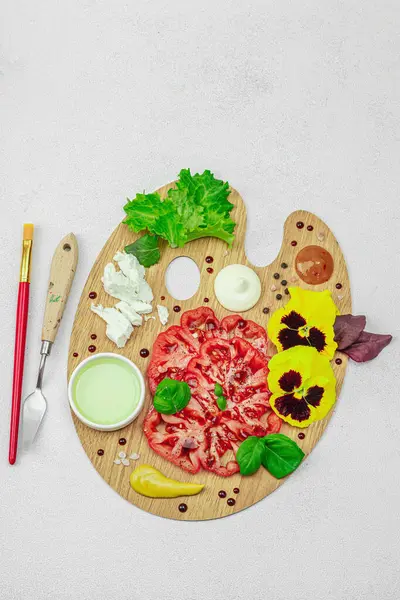 Dilimlenmiş aile yadigarı domates, feta, otlar, yenilebilir çiçekler, ve ahşap palette soslar, fırça ve palet bıçağıyla şekillendirilmiş. Hafif taş arkaplan, üst görünüm