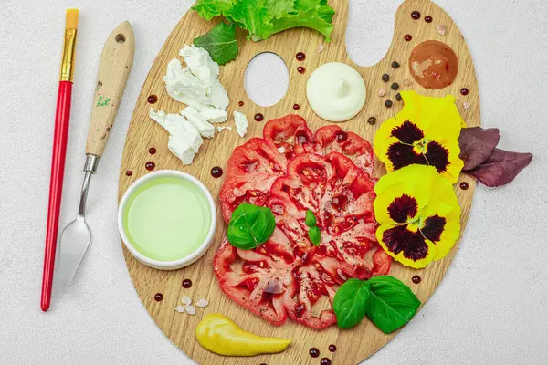 Dilimlenmiş aile yadigarı domates, feta, otlar, yenilebilir çiçekler, ve ahşap palette soslar, fırça ve palet bıçağıyla şekillendirilmiş. Hafif taş arkaplan, üst görünüm