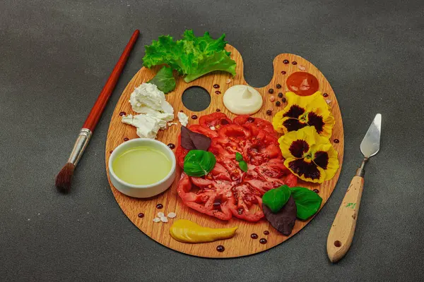 Dilimlenmiş aile yadigarı domates, feta, otlar, yenilebilir çiçekler, ve ahşap palette soslar, fırça ve palet bıçağıyla şekillendirilmiş. Koyu taş arkaplan, kopyalama alanı