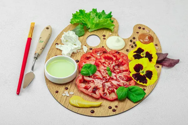 Dilimlenmiş aile yadigarı domates, feta, otlar, yenilebilir çiçekler, ve ahşap palette soslar, fırça ve palet bıçağıyla şekillendirilmiş. Hafif taş arkaplan, kapat