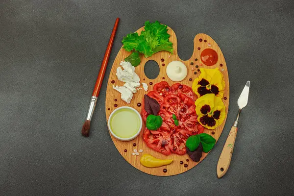 Dilimlenmiş aile yadigarı domates, feta, otlar, yenilebilir çiçekler, ve ahşap palette soslar, fırça ve palet bıçağıyla şekillendirilmiş. Koyu taş zemin, üst görünüm