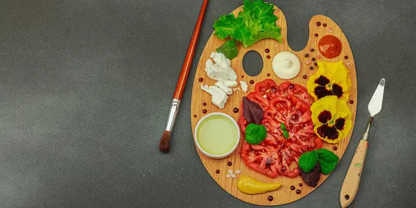 Dilimlenmiş aile yadigarı domates, feta, otlar, yenilebilir çiçekler, ve ahşap palette soslar, fırça ve palet bıçağıyla şekillendirilmiş. Koyu taş arkaplan, pankart biçimi