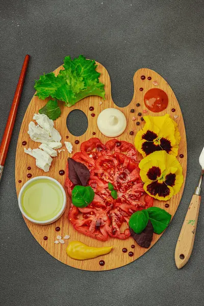 Dilimlenmiş aile yadigarı domates, feta, otlar, yenilebilir çiçekler, ve ahşap palette soslar, fırça ve palet bıçağıyla şekillendirilmiş. Koyu taş zemin, üst görünüm