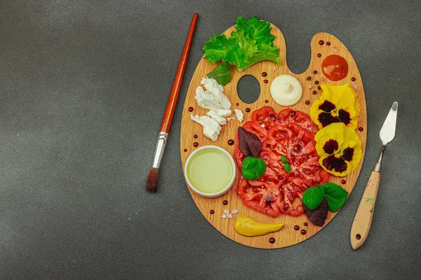 Dilimlenmiş aile yadigarı domates, feta, otlar, yenilebilir çiçekler, ve ahşap palette soslar, fırça ve palet bıçağıyla şekillendirilmiş. Koyu taş zemin, üst görünüm