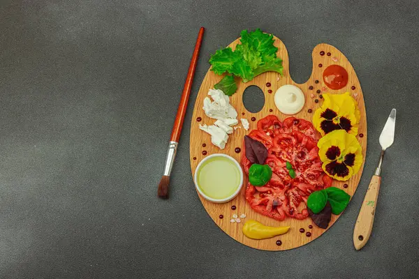 Dilimlenmiş aile yadigarı domates, feta, otlar, yenilebilir çiçekler, ve ahşap palette soslar, fırça ve palet bıçağıyla şekillendirilmiş. Koyu taş zemin, üst görünüm