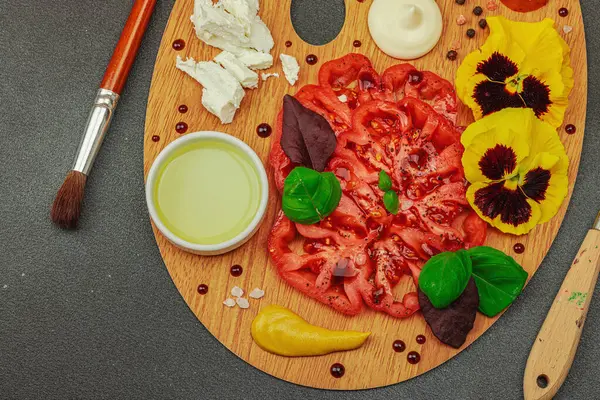 Dilimlenmiş aile yadigarı domates, feta, otlar, yenilebilir çiçekler, ve ahşap palette soslar, fırça ve palet bıçağıyla şekillendirilmiş. Koyu taş zemin, üst görünüm