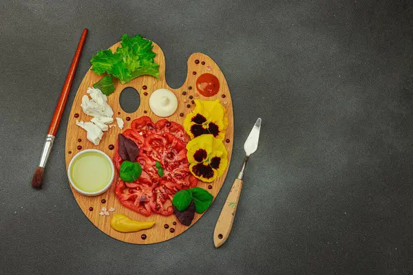 Dilimlenmiş aile yadigarı domates, feta, otlar, yenilebilir çiçekler, ve ahşap palette soslar, fırça ve palet bıçağıyla şekillendirilmiş. Koyu taş zemin, üst görünüm