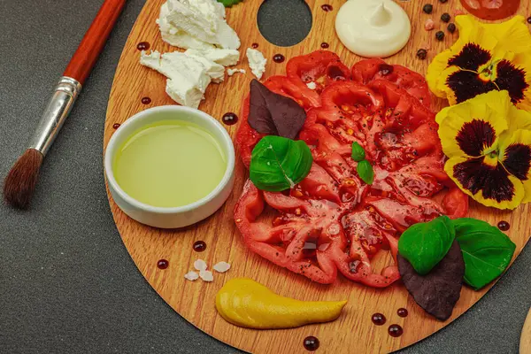 Dilimlenmiş aile yadigarı domates, feta, otlar, yenilebilir çiçekler, ve ahşap palette soslar, fırça ve palet bıçağıyla şekillendirilmiş. Koyu taş beton arka plan, kapat