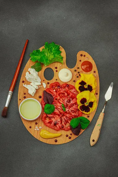 Dilimlenmiş aile yadigarı domates, feta, otlar, yenilebilir çiçekler, ve ahşap palette soslar, fırça ve palet bıçağıyla şekillendirilmiş. Koyu taş zemin, üst görünüm