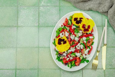 Domatesli taze vegan salatası, roka, keçi peyniri, zeytin ve beyaz tabakta yenilebilir çiçekler. Çevre dostu ve estetik titreşimli sağlıklı yemek konsepti. Moda kiremitleri arka plan, üst görünüm