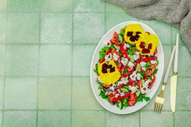 Domatesli taze vegan salatası, roka, keçi peyniri, zeytin ve beyaz tabakta yenilebilir çiçekler. Çevre dostu ve estetik titreşimli sağlıklı yemek konsepti. Moda kiremitleri arka plan, üst görünüm