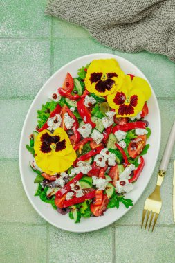 Domatesli taze vegan salatası, roka, keçi peyniri, zeytin ve beyaz tabakta yenilebilir çiçekler. Çevre dostu ve estetik titreşimli sağlıklı yemek konsepti. Moda kiremitleri arka plan, üst görünüm