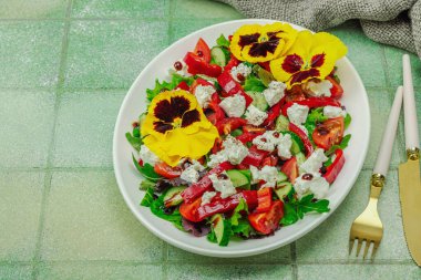 Domatesli taze vegan salatası, roka, keçi peyniri, zeytin ve beyaz tabakta yenilebilir çiçekler. Çevre dostu ve estetik titreşimli sağlıklı yemek konsepti. Moda kiremitleri arka planı, kopyalama alanı