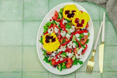 Domatesli taze vegan salatası, roka, keçi peyniri, zeytin ve beyaz tabakta yenilebilir çiçekler. Çevre dostu ve estetik titreşimli sağlıklı yemek konsepti. Moda kiremitleri arka plan, üst görünüm
