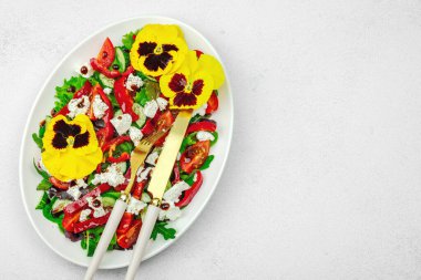 Domatesli taze vegan salatası, roka, keçi peyniri, zeytin ve beyaz tabakta yenilebilir çiçekler. Çevre dostu ve estetik titreşimli sağlıklı yemek konsepti. Beyaz arkaplan, üst görünüm
