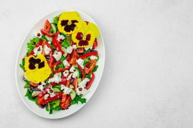 Domatesli taze vegan salatası, roka, keçi peyniri, zeytin ve beyaz tabakta yenilebilir çiçekler. Çevre dostu ve estetik titreşimli sağlıklı yemek konsepti. Beyaz arkaplan, üst görünüm