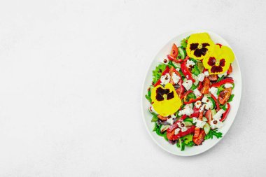 Domatesli taze vegan salatası, roka, keçi peyniri, zeytin ve beyaz tabakta yenilebilir çiçekler. Çevre dostu ve estetik titreşimli sağlıklı yemek konsepti. Beyaz arkaplan, üst görünüm