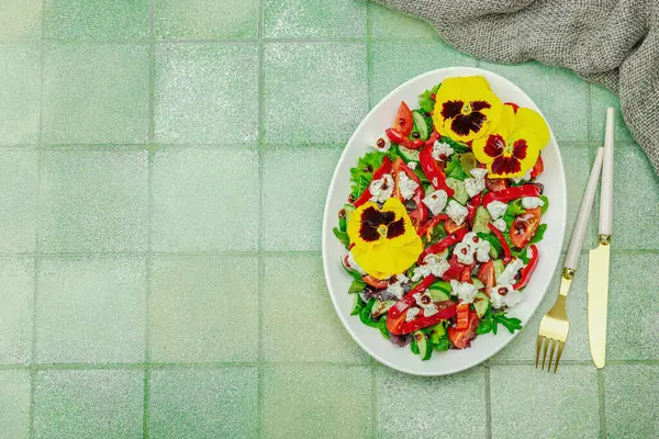 Domatesli taze vegan salatası, roka, keçi peyniri, zeytin ve beyaz tabakta yenilebilir çiçekler. Çevre dostu ve estetik titreşimli sağlıklı yemek konsepti. Moda kiremitleri arka plan, üst görünüm