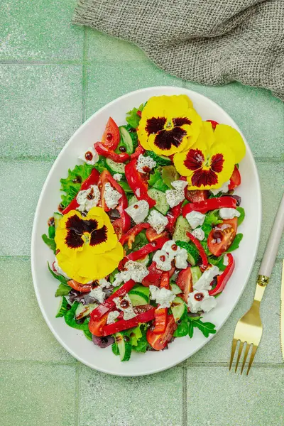 Domatesli taze vegan salatası, roka, keçi peyniri, zeytin ve beyaz tabakta yenilebilir çiçekler. Çevre dostu ve estetik titreşimli sağlıklı yemek konsepti. Moda kiremitleri arka plan, üst görünüm