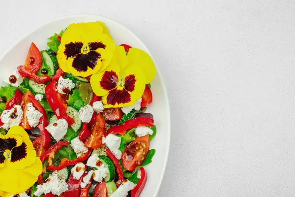 Domatesli taze vegan salatası, roka, keçi peyniri, zeytin ve beyaz tabakta yenilebilir çiçekler. Çevre dostu ve estetik titreşimli sağlıklı yemek konsepti. Beyaz arkaplan, üst görünüm
