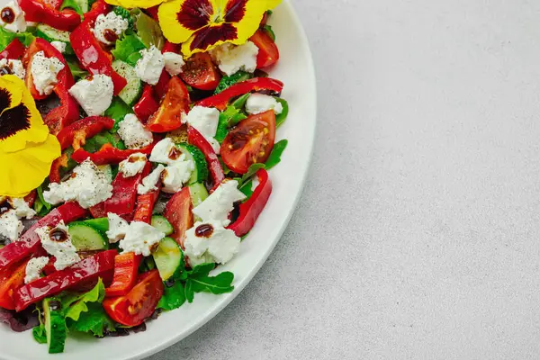 Domatesli taze vegan salatası, roka, keçi peyniri, zeytin ve beyaz tabakta yenilebilir çiçekler. Çevre dostu ve estetik titreşimli sağlıklı yemek konsepti. Beyaz arkaplan, kapat