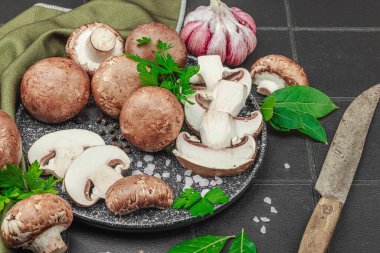 Sarımsaklı, otlu taze kahverengi mantarlar ve siyah fayanslı mutfak yüzeyinde kırsal bıçak. Moda seramik plaka, düz döşeme, yakın çekim.