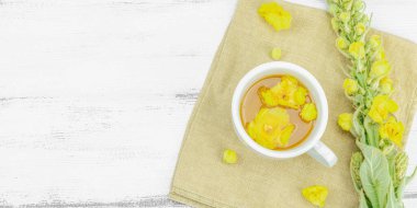 Tarafsız ahşap arka planda yaprakları ve narin çiçekleri olan bir fincan bitki çayı. Doğal ışık, alternatif tıp, klasik şifalı bitki havası, afiş biçimi