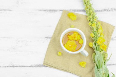 Tarafsız ahşap arka planda yaprakları ve narin çiçekleri olan bir fincan bitki çayı. Doğal ışık, alternatif tıp, klasik şifalı bitki havası, üst görünüm.