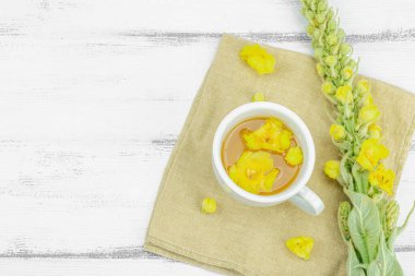 Tarafsız ahşap arka planda yaprakları ve narin çiçekleri olan bir fincan bitki çayı. Doğal ışık, alternatif tıp, klasik şifalı bitki havası, üst görünüm.