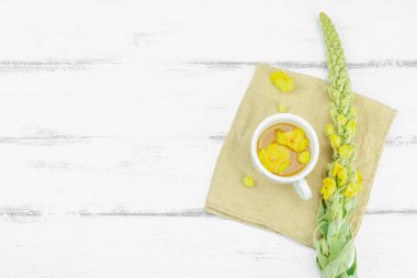 Tarafsız ahşap arka planda yaprakları ve narin çiçekleri olan bir fincan bitki çayı. Doğal ışık, alternatif tıp, klasik şifalı bitki havası, üst görünüm.