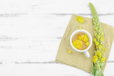 Tarafsız ahşap arka planda yaprakları ve narin çiçekleri olan bir fincan bitki çayı. Doğal ışık, alternatif tıp, klasik şifalı bitki havası, üst görünüm.