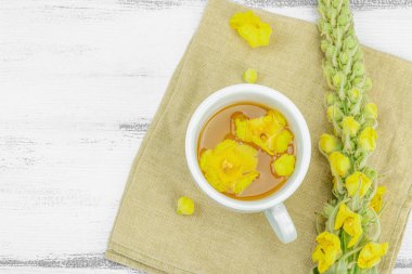 Tarafsız ahşap arka planda yaprakları ve narin çiçekleri olan bir fincan bitki çayı. Doğal ışık, alternatif tıp, klasik şifalı bitki havası, üst görünüm.