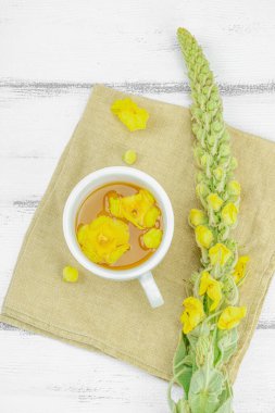 Tarafsız ahşap arka planda yaprakları ve narin çiçekleri olan bir fincan bitki çayı. Doğal ışık, alternatif tıp, klasik şifalı bitki havası, üst görünüm.