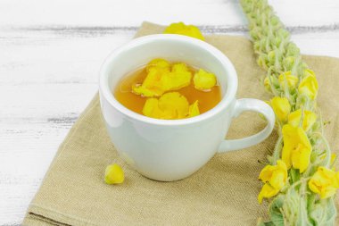 Tarafsız ahşap arka planda yaprakları ve narin çiçekleri olan bir fincan bitki çayı. Doğal ışık, alternatif tıp, klasik şifalı bitki havası, kapat.