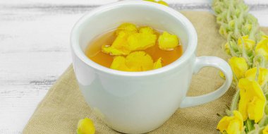 Tarafsız ahşap arka planda yaprakları ve narin çiçekleri olan bir fincan bitki çayı. Doğal ışık, alternatif tıp, klasik şifalı bitki havası, afiş biçimi