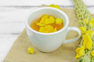 Tarafsız ahşap arka planda yaprakları ve narin çiçekleri olan bir fincan bitki çayı. Doğal ışık, alternatif tıp, klasik şifalı bitki havası, kapat.