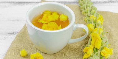 Tarafsız ahşap arka planda yaprakları ve narin çiçekleri olan bir fincan bitki çayı. Doğal ışık, alternatif tıp, klasik şifalı bitki havası, afiş biçimi