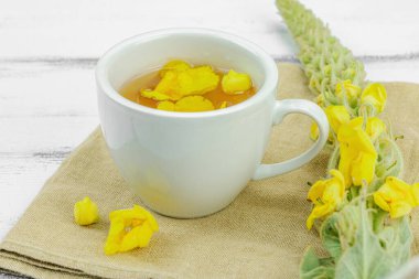 Tarafsız ahşap arka planda yaprakları ve narin çiçekleri olan bir fincan bitki çayı. Doğal ışık, alternatif tıp, klasik şifalı bitki havası, kapat.