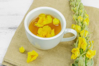 Tarafsız ahşap arka planda yaprakları ve narin çiçekleri olan bir fincan bitki çayı. Doğal ışık, alternatif tıp, klasik şifalı bitki havası, kapat.