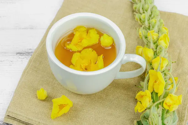 Tarafsız ahşap arka planda yaprakları ve narin çiçekleri olan bir fincan bitki çayı. Doğal ışık, alternatif tıp, klasik şifalı bitki havası, kapat.