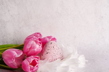 Easter egg in white nest of feathers with pink tulips on white concrete background