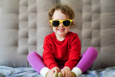 Kırmızı elbiseli, sarı gözlüklü, gülümseyen ve evdeki kanepede oturan küçük güzel bir kız.