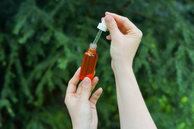 Kırmızı cam şişeyi kaplıca yağıyla tutan kadın elleri. Salon terapisi, doğal eko vücut bakım ürünleri, yeşil doğa geçmişi