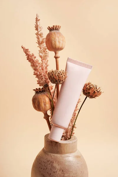 Cosmetic Cream pink tube mockup with autumn flowers in the wooden vase on beige background. Natural skin care beauty product concept