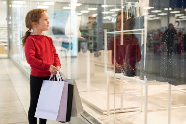 Kırmızı kazaklı, elbise dükkanında torbaları olan tatlı bir kız çocuğu.