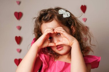 Pembe elbiseli şirin, gülümseyen küçük kız kalp şeklinde jest yapıyor.